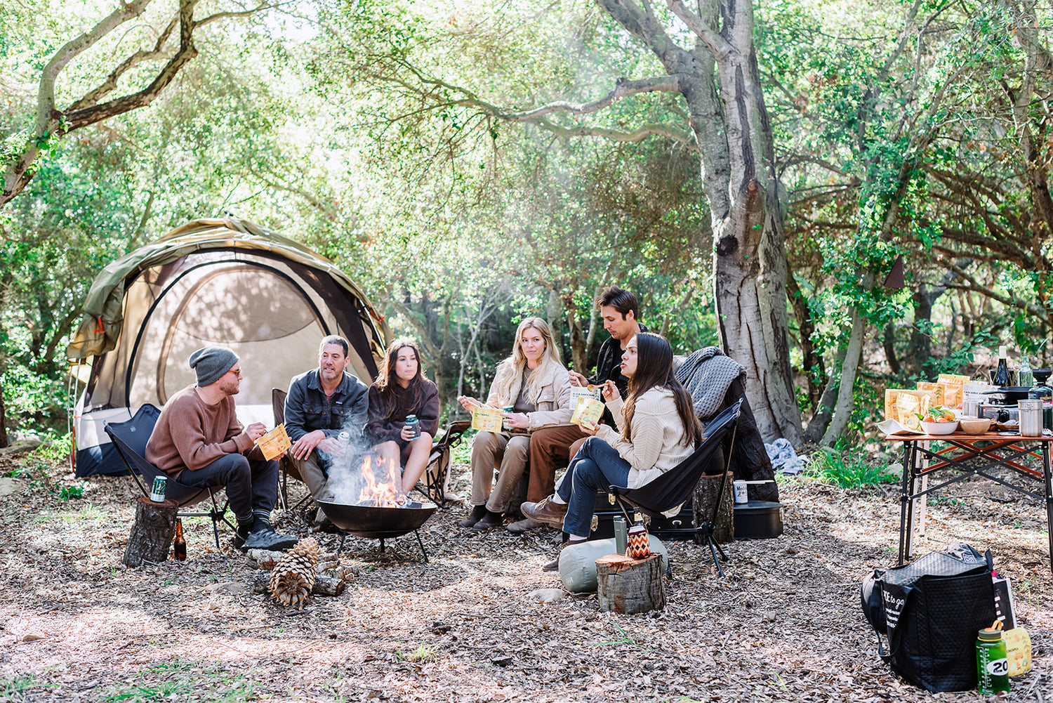 Campers huddles around a campfire, eating vegan camping meals by poe and co. folk foods